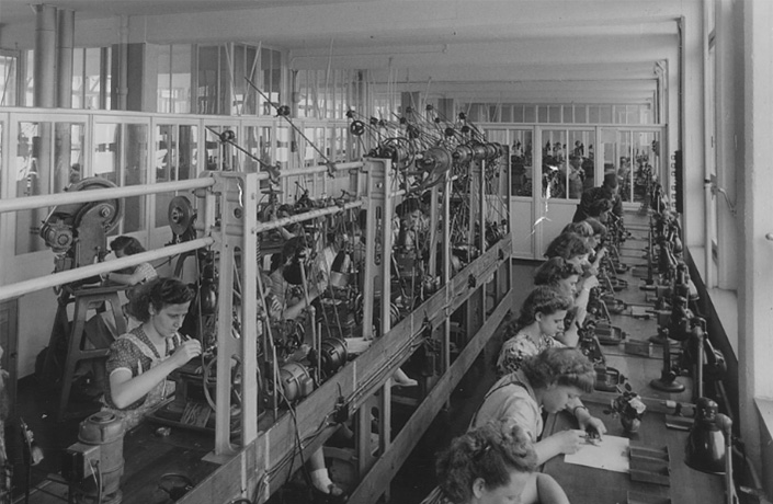 Schwarzweißaufnahme einer Fertigungshalle mit Frauen an Maschinen – historische Aufnahme der Firma.