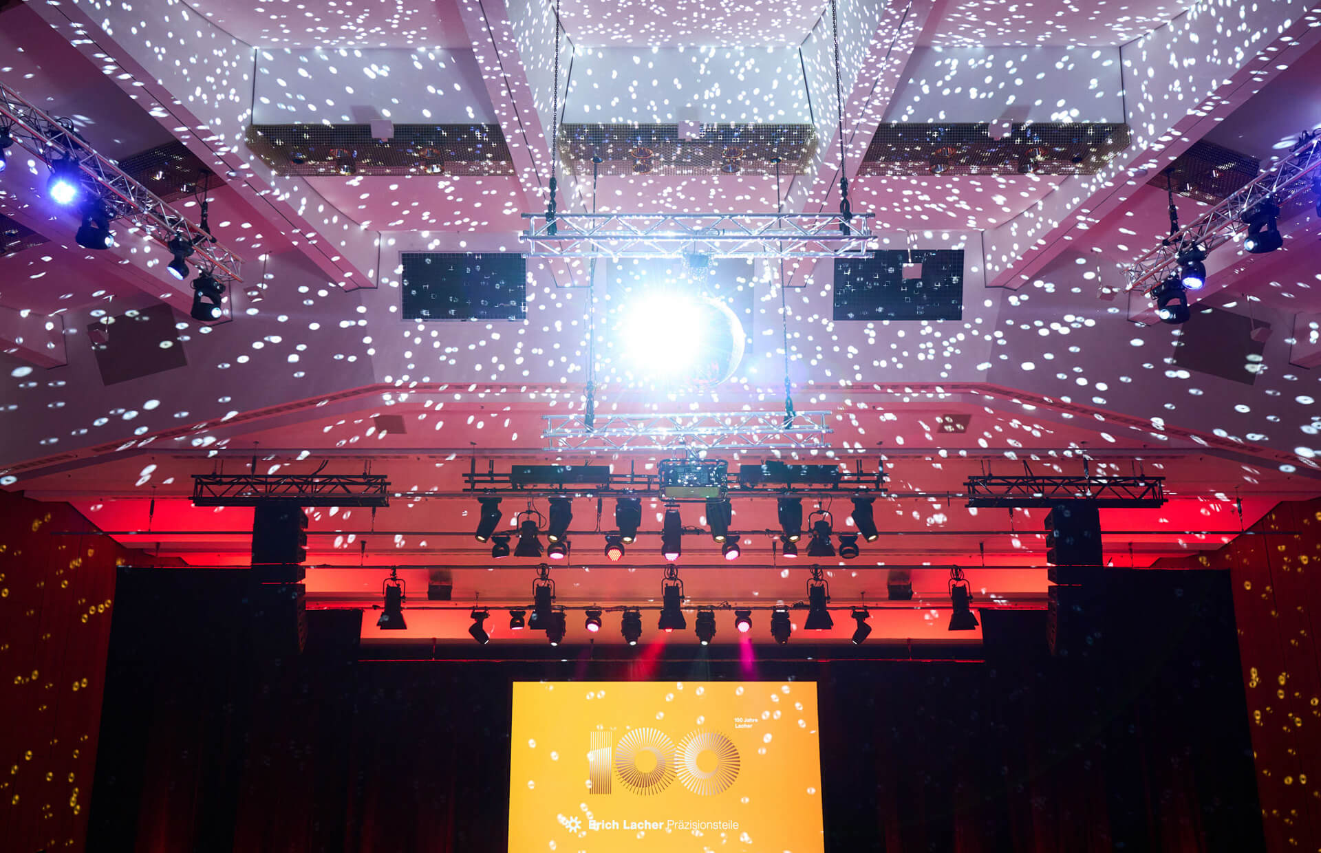 Festlich beleuchteter Veranstaltungssaal mit Lichtprojektionen an der Decke und Bühne – im Zentrum ein leuchtender Screen mit dem Schriftzug ‚100 Jahre Erich Lacher Präzisionsteile‘.