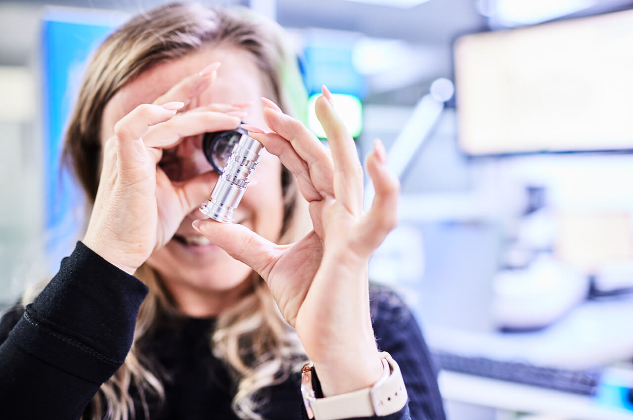 Mitarbeiterin inspiziert Präzisionsteil mit Lupe in der visuellen Qualitätsprüfung im Messraum.