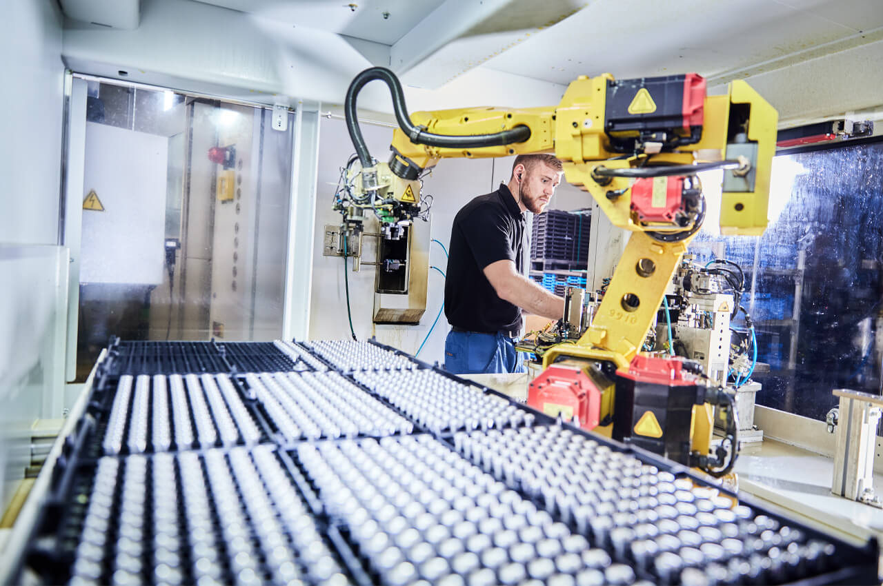 Roboterarm bestückt Produktionsanlage mit Bauteilen, Mitarbeiter überwacht den automatisierten Ablauf.