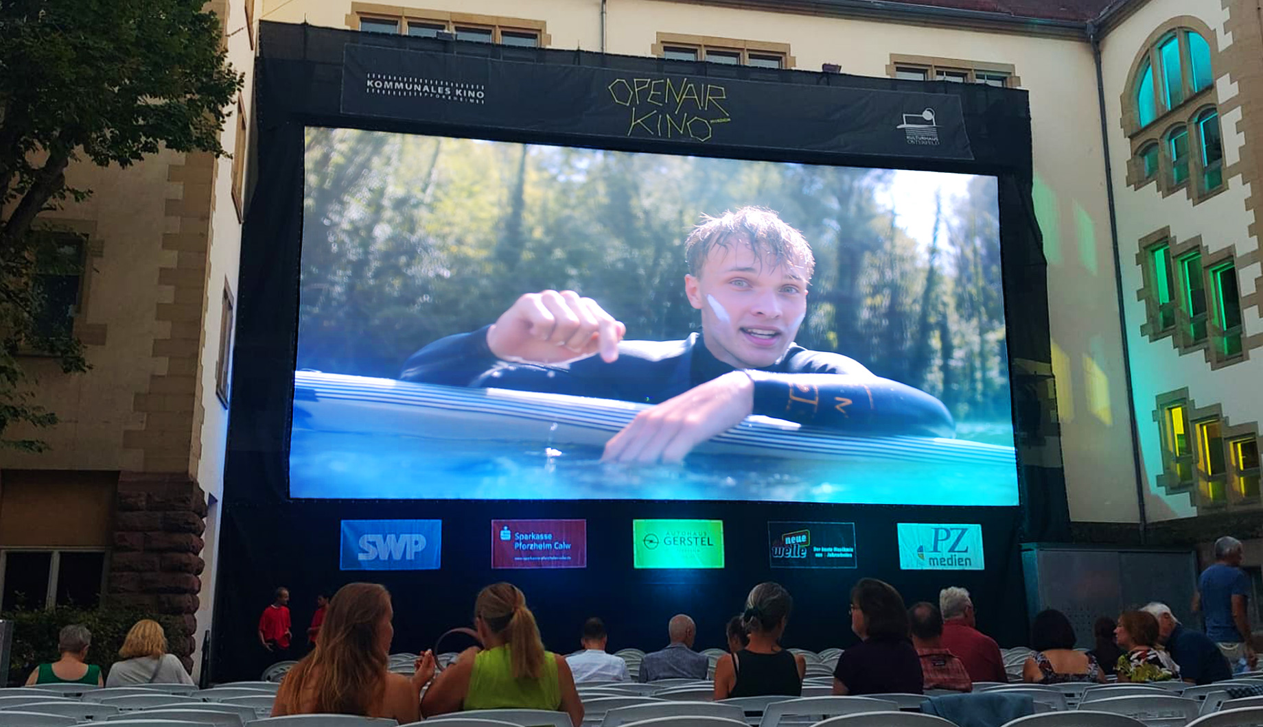 Große Leinwand im Open-Air-Kino zeigt Surfer im Wasser, Publikum sitzt davor.