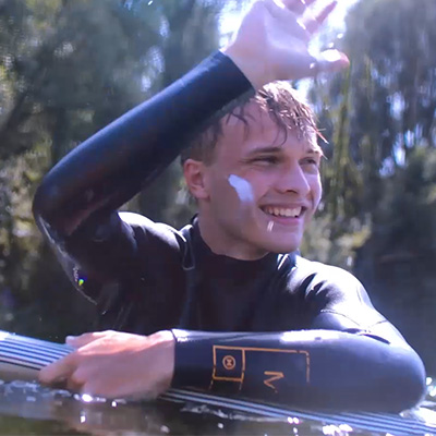 Junger Mann im Neoprenanzug im Fluss, lächelnd mit Surfboard im Arm.