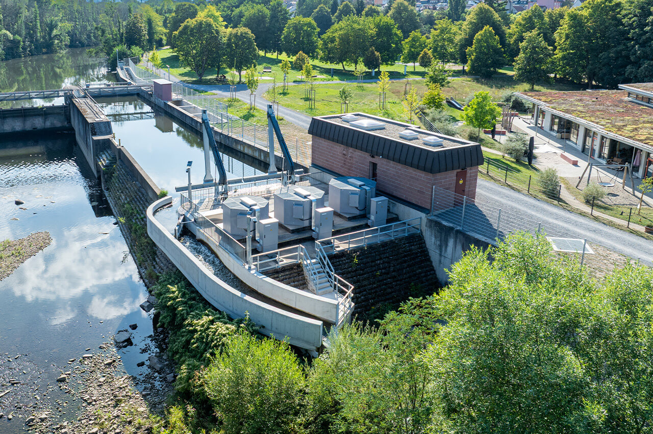 Wasserkraftwerk an einem Fluss mit Wehr, Gebäuden und Grünflächen.