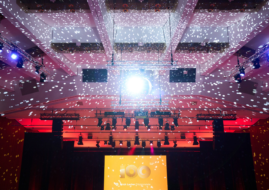 Festlich beleuchteter Veranstaltungssaal mit Lichtprojektionen an der Decke und Bühne – im Zentrum ein leuchtender Screen mit dem Schriftzug ‚100 Jahre Erich Lacher Präzisionsteile‘.