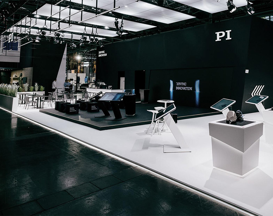Großer schwarzer Hightech-Messestand von PI mit beleuchtetem Claim „Driving Innovation“, interaktiven Touchscreens, Präsentationsinseln und klarer Zonierung in Weiß und Schwarz.