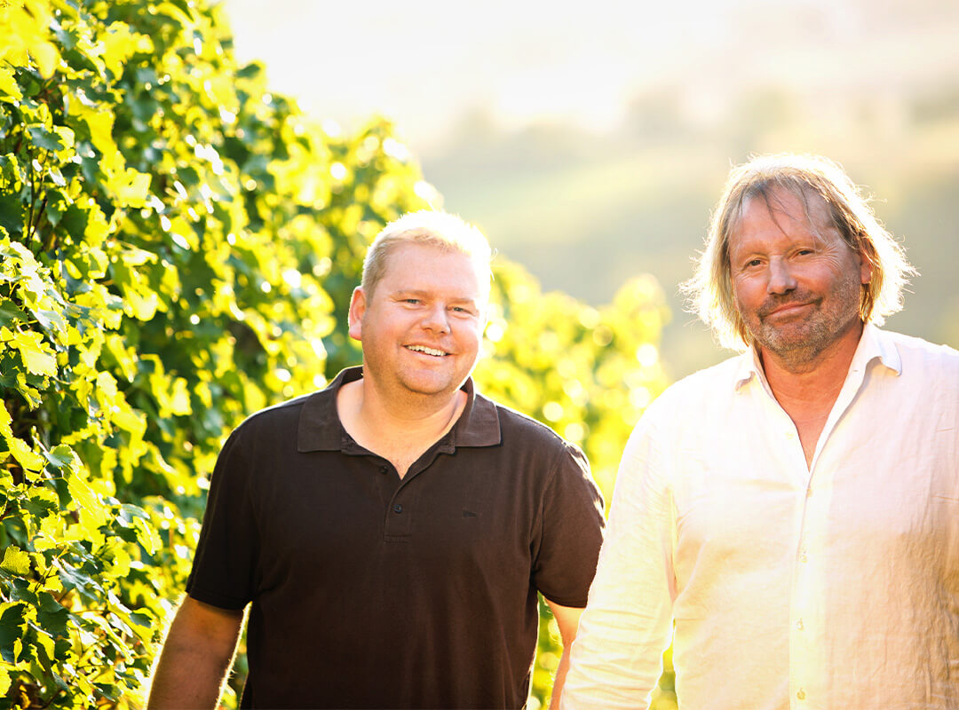 Claus und Robin Bischoff stehen freundlich lächelnd vor Weinreben.