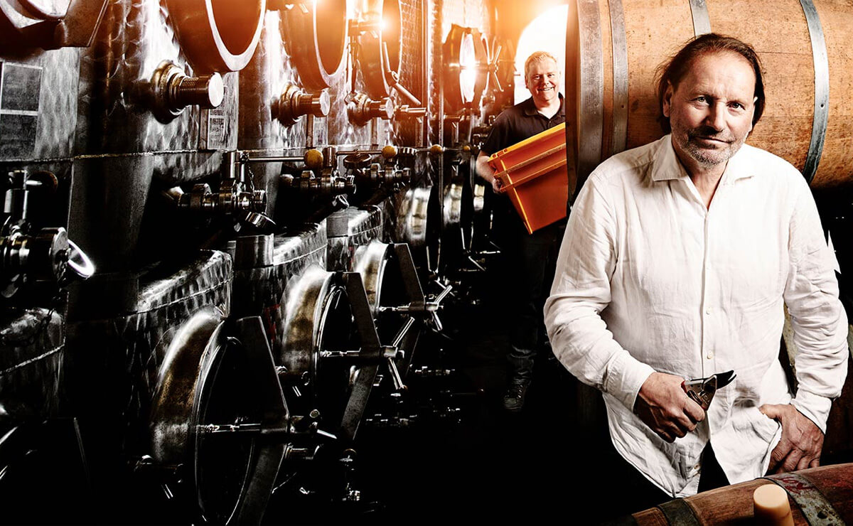Mann im weißen Hemd im Weinkeller neben Holzfässern. Dahinter Mann in Schwarz mit orangenen Wannen.